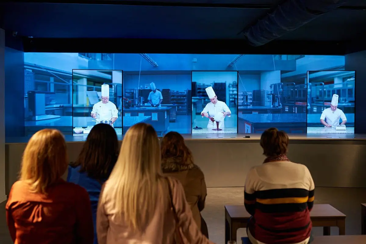 Plusieurs personnes sont assises face à un grand écran montrant trois images d'un chef dans une cuisine.