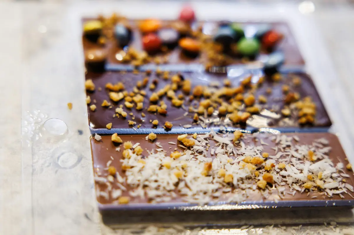Gros plan sur une barre de chocolat divisée en plusieurs parties, garnie de noix concassées et de morceaux de bonbons colorés.