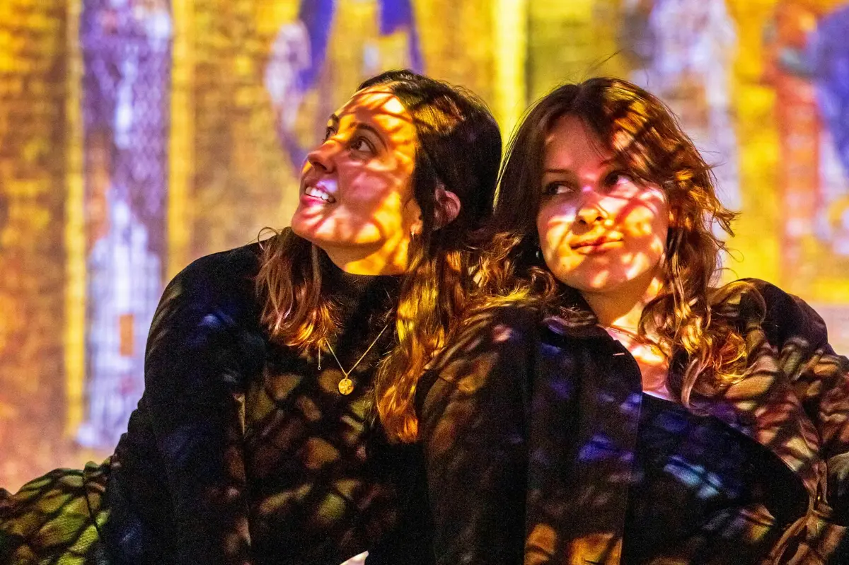 Deux femmes sur lesquelles sont projetés des motifs lumineux colorés, assises sur un fond flou aux teintes vibrantes.