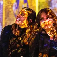 Deux femmes sur lesquelles sont projetés des motifs lumineux colorés, assises sur un fond flou aux teintes vibrantes. &copy; ®Cités Immersives