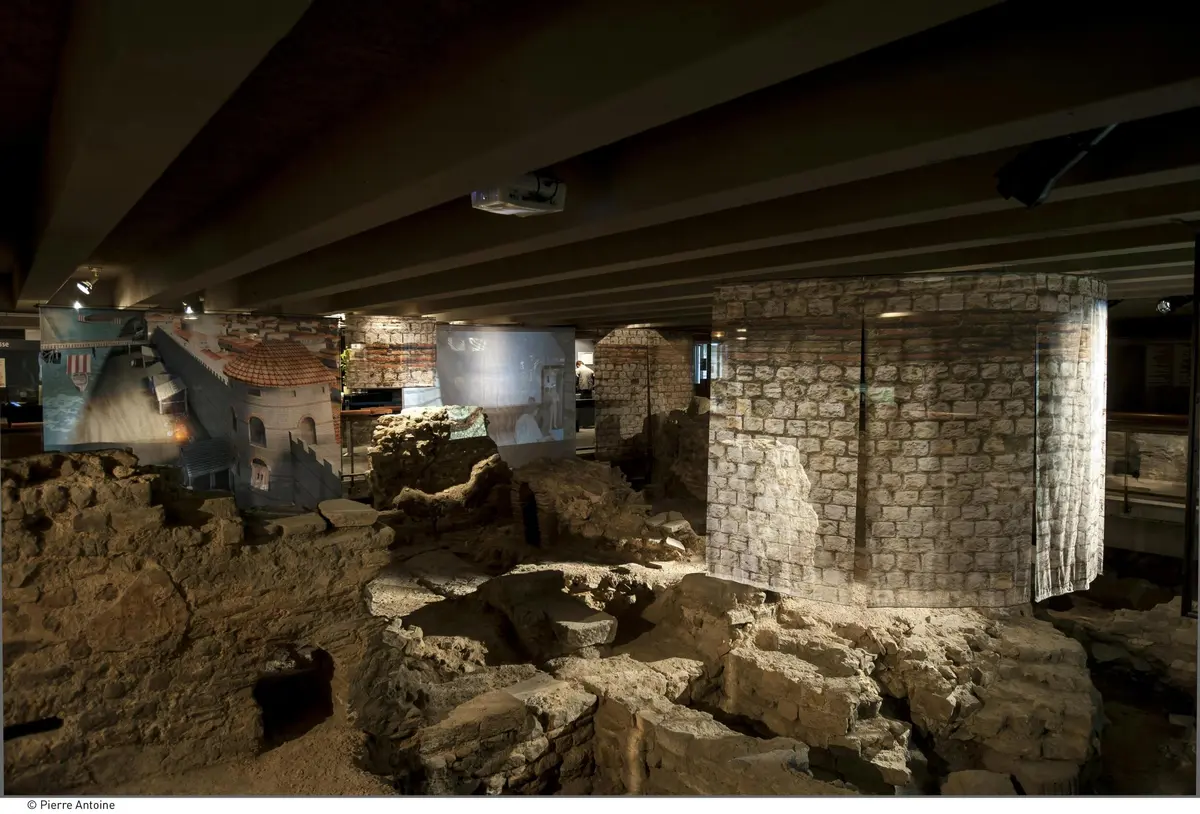 Anciennes ruines en pierre sous un bâtiment aux murs de briques et aux poutres en bois, avec des piliers et des images d'anciennes structures projetées sur les murs.