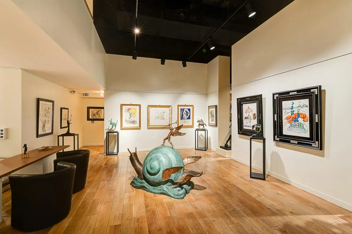 Galerie d'art avec des tableaux encadrés sur des murs blancs, sol en bois, plafond noir, sculpture d'un escargot avec une figure humaine au centre.