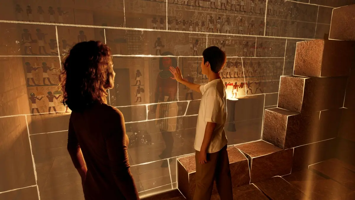 Un homme touche une paroi en verre exposant des peintures égyptiennes anciennes, tandis qu'une femme observe la scène. Des blocs de pierre et une lampe se trouvent à l'arrière-plan.