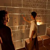 Un homme touche une paroi en verre exposant des peintures égyptiennes anciennes, tandis qu'une femme observe la scène. Des blocs de pierre et une lampe se trouvent à l'arrière-plan. &copy; TDB