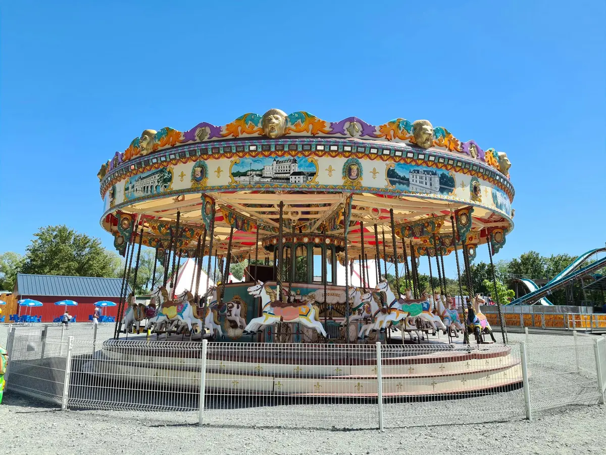 Un carrousel coloré et orné de chevaux peints est entouré d'une clôture blanche sous un ciel bleu clair.