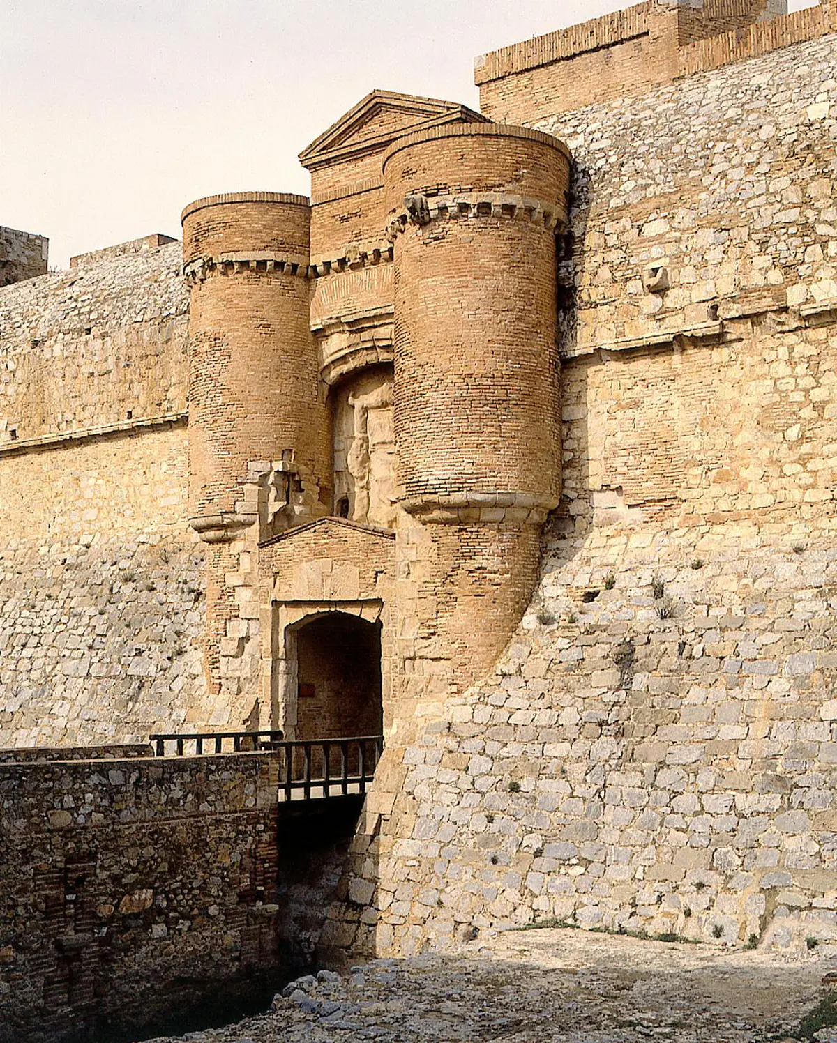 Forteresse en pierre avec deux grandes tours cylindriques et une porte. La structure est faite de pierres brutes et de briques.