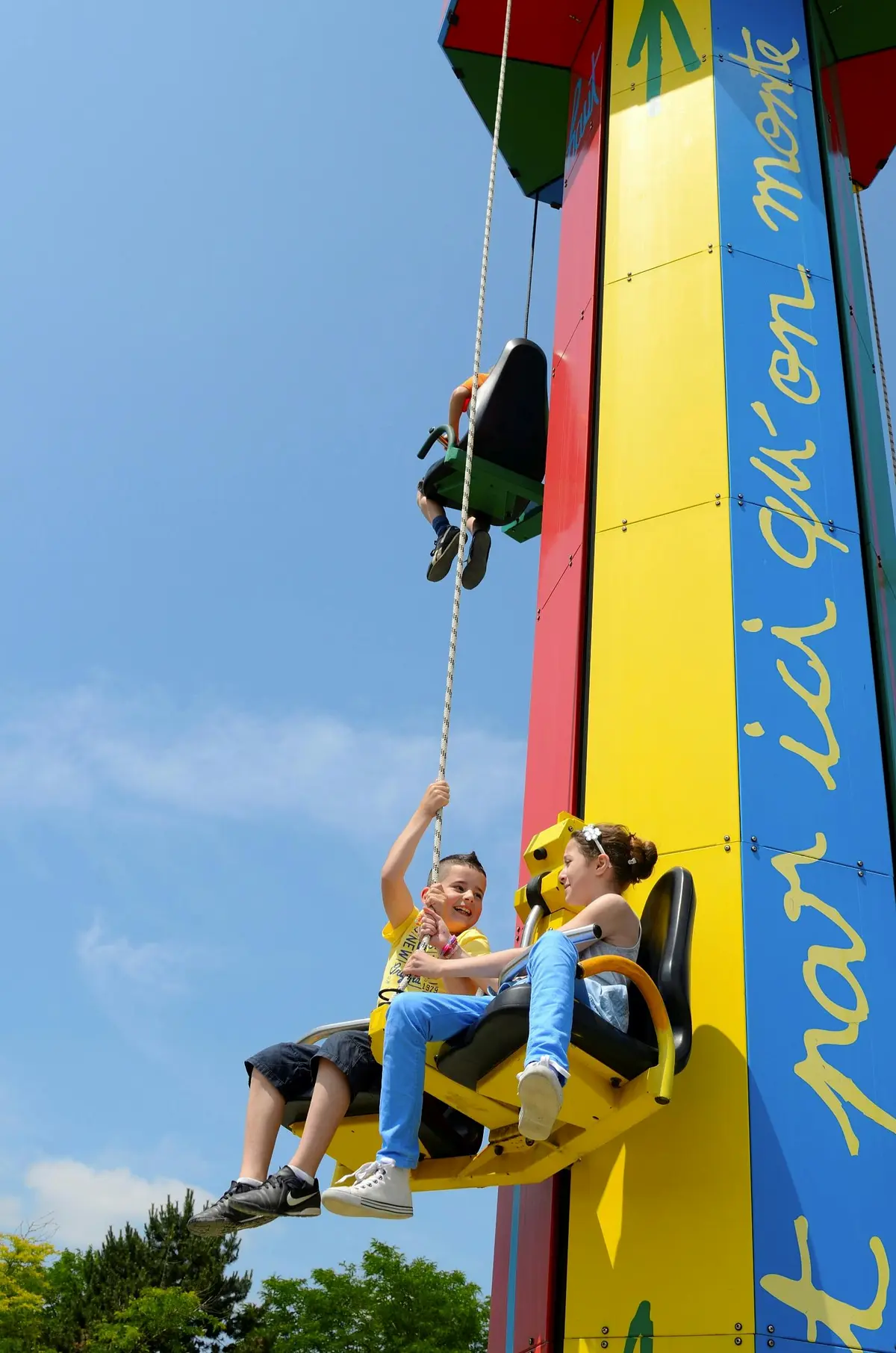 Deux enfants souriant sur un siège de balançoire montant un manège vertical coloré dans un ciel bleu clair. Des arbres sont visibles à l'arrière-plan.