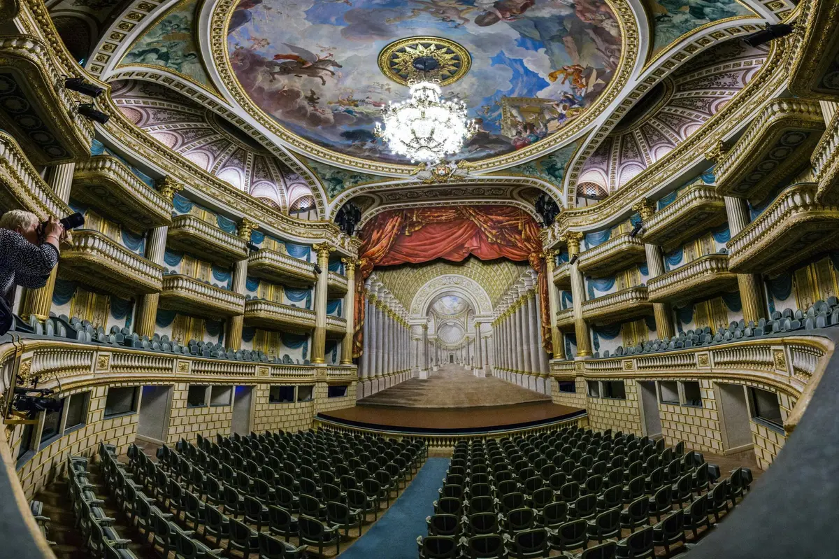 Un théâtre somptueux, avec un immense lustre, un plafond peint, un rideau rouge, une scène et des rangées de sièges vides.