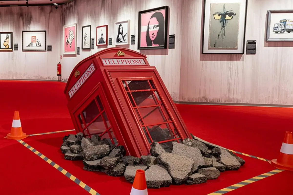 Une cabine téléphonique britannique rouge partiellement enfouie sous les décombres, dans un espace d'exposition où sont accrochées des œuvres d'art encadrées aux murs.