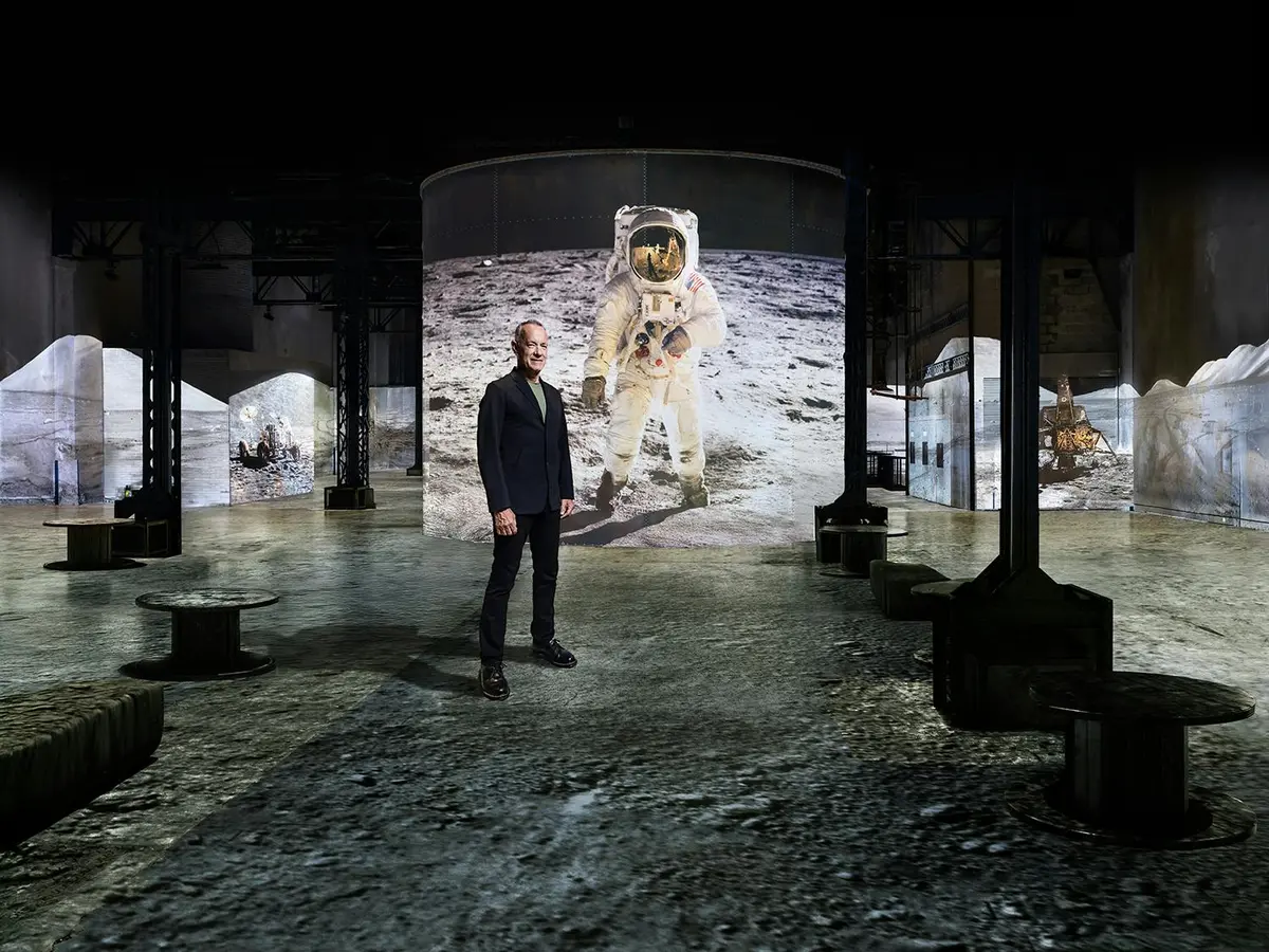 Un homme se tient dans un espace d'exposition où sont projetées de grandes images d'un astronaute sur la lune.