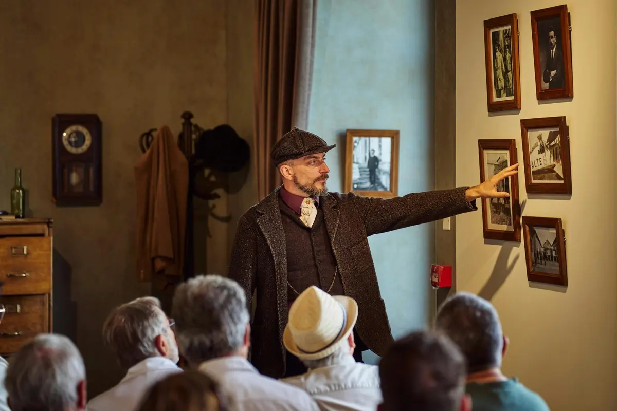 Un homme vêtu d'une tenue d'époque s'adresse à un public, en montrant du doigt des photos encadrées accrochées au mur d'une pièce faiblement éclairée.