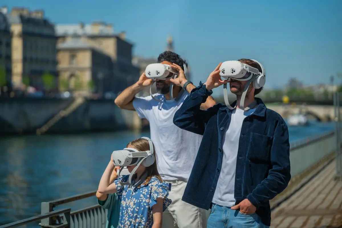 Trois personnes, deux adultes et un enfant, portent des casques de réalité virtuelle en plein air, près d'un plan d'eau et d'un bâtiment historique.