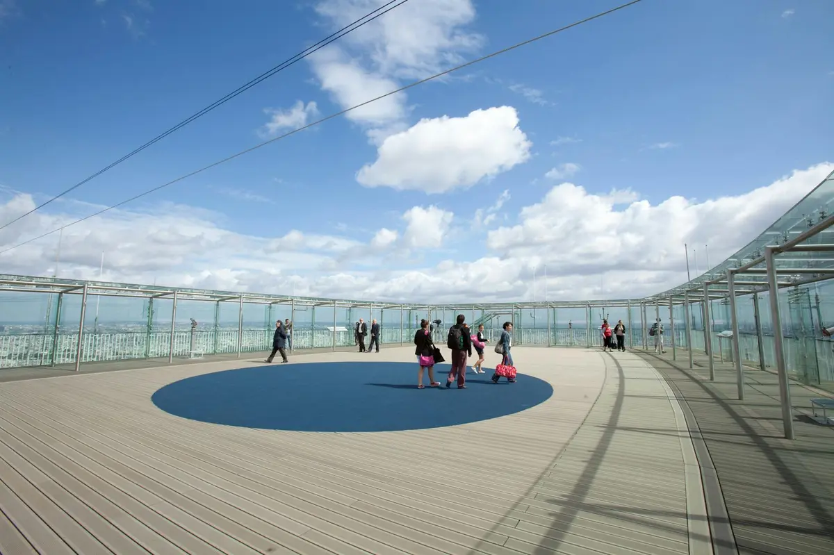 Les gens marchent et se tiennent debout sur un grand pont d'observation en plein air avec un centre circulaire bleu sous un ciel partiellement nuageux.