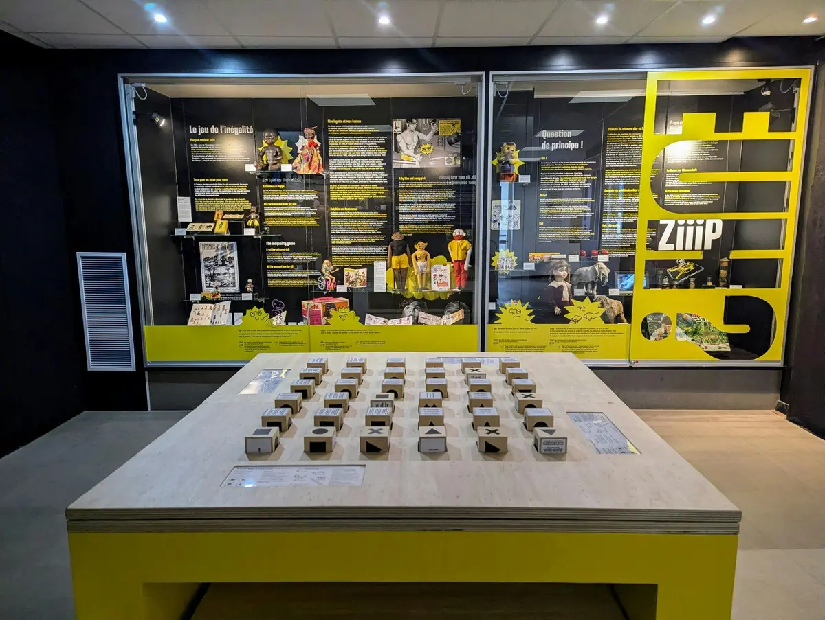 Exposition avec des panneaux de texte, des accents jaunes et des pièces de jeu interactives sur une table, le tout à l'intérieur d'une salle d'exposition.