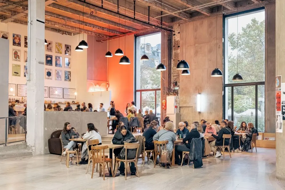 Un café animé avec des personnes assises à des tables, de grandes fenêtres, des lampes suspendues et un décor industriel moderne.