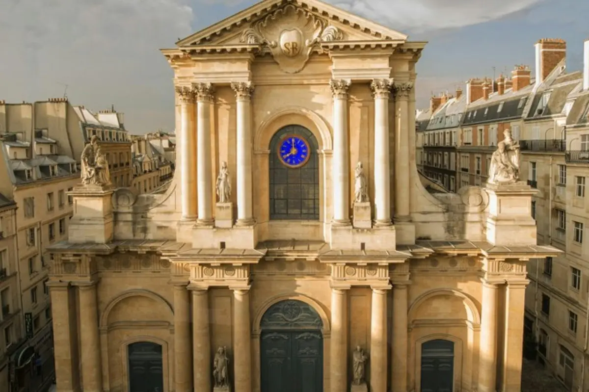 Façade d'un bâtiment historique richement décoré, avec des colonnes, des statues et une fenêtre circulaire bleue, sur fond de paysage urbain sous un ciel nuageux.