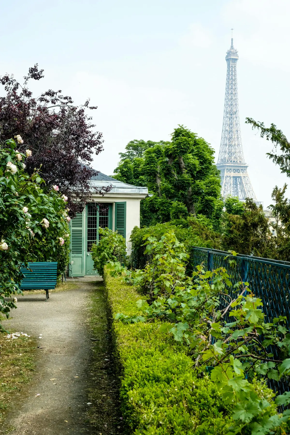 Allée menant à un petit bâtiment aux volets verts, entouré de verdure et de fleurs, avec la Tour Eiffel en arrière-plan.
