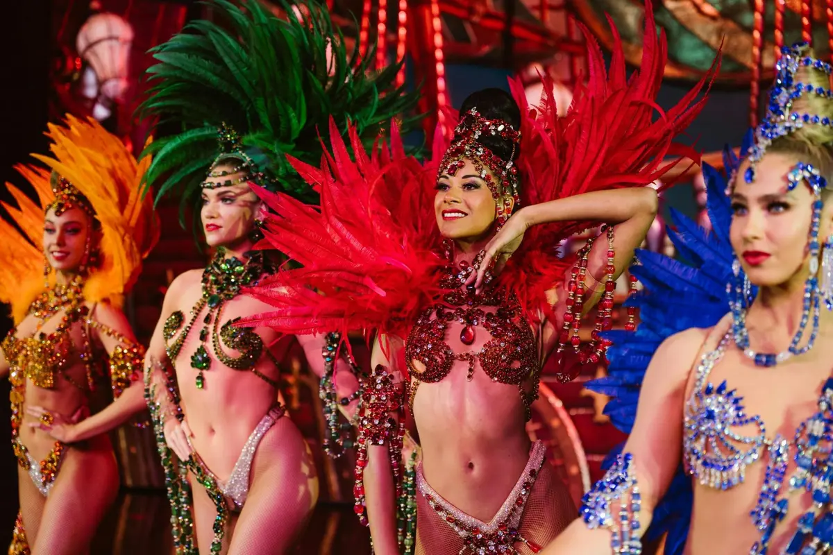 Des artistes vêtus de costumes somptueux ornés de plumes, aux nuances de rouge, de vert et de bleu, dansent sur une scène aux couleurs éclatantes.