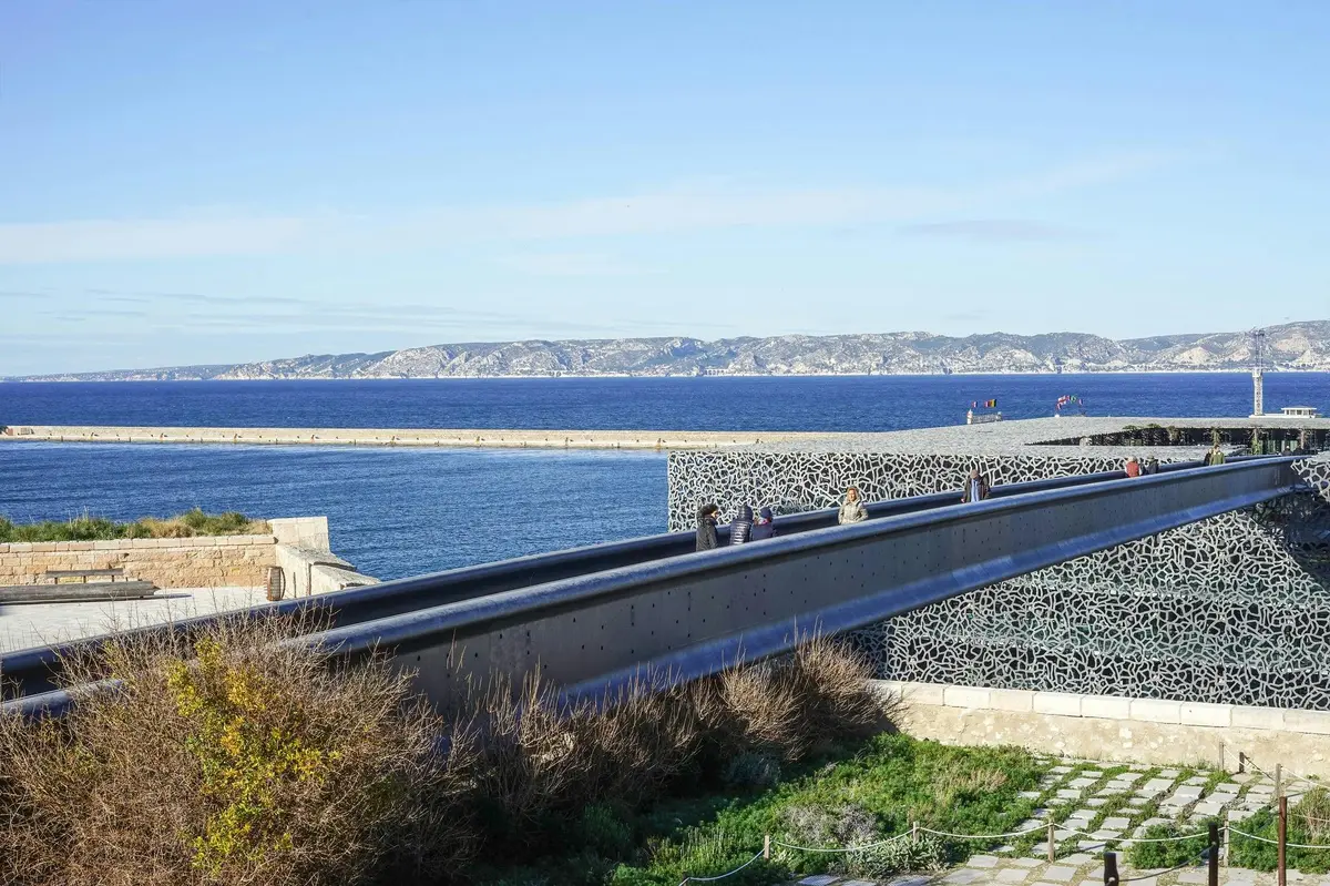Des personnes marchent sur un pont aux côtés en treillis complexes, surplombant la mer et les montagnes lointaines sous un ciel bleu clair.