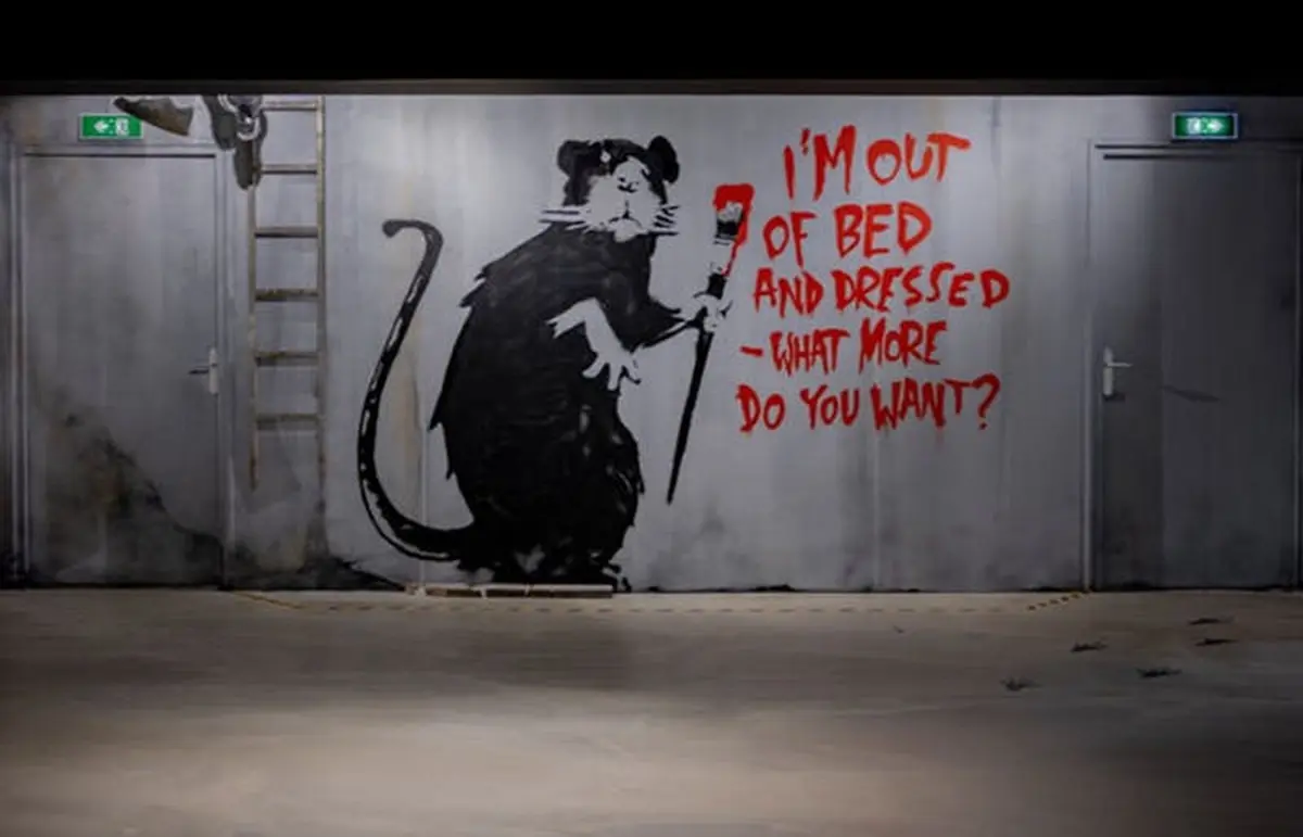 Graffiti d'un gros rat tenant un pinceau avec de la peinture rouge, à côté d'un texte indiquant I'M OUT OF BED AND DRESSED -WHAT MORE DO YOU WANT ? (Je suis sorti du lit et habillé - que voulez-vous de plus ?).