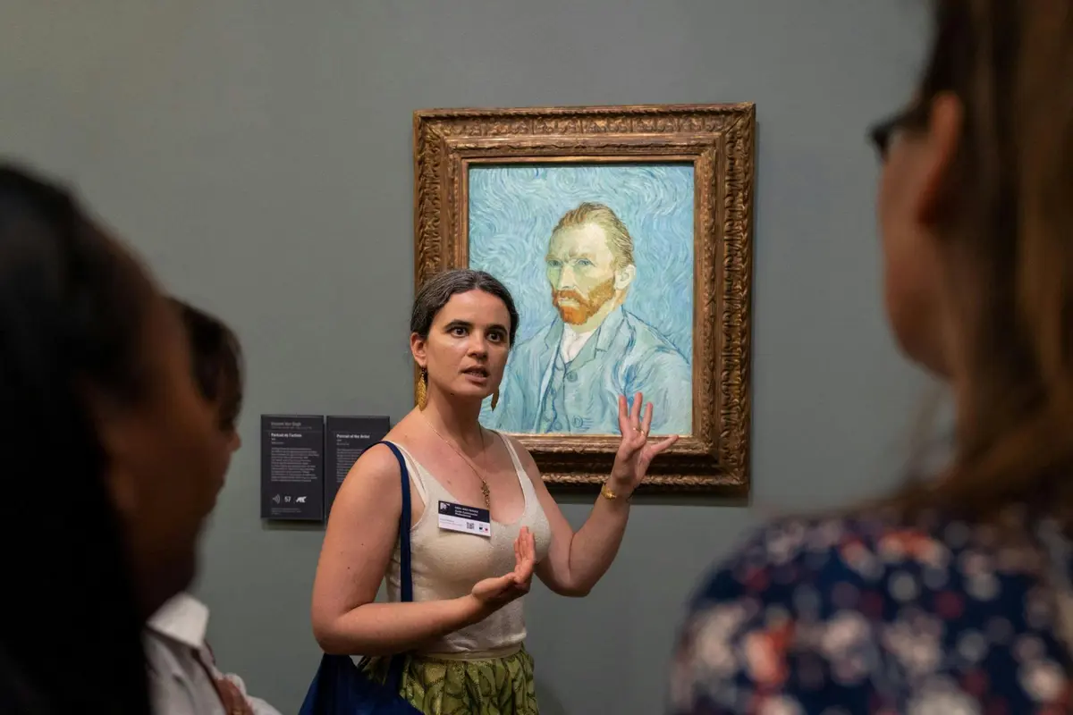 Une personne parle en faisant des gestes près d'un tableau encadré représentant un homme à la barbe rouge sur fond bleu dans un musée.