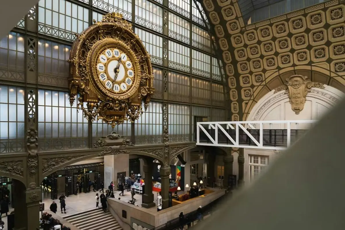Une horloge richement décorée est accrochée dans un intérieur spacieux, avec de hautes fenêtres, des éléments architecturaux décoratifs et des passants qui défilent en contrebas.
