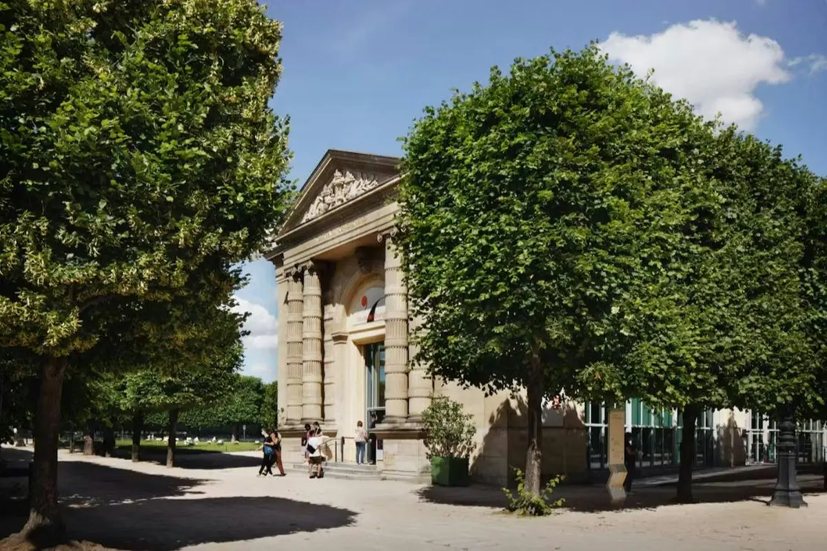 Un bâtiment de style classique, avec des colonnes et un fronton, entouré d'arbres, avec des passants qui se promènent à proximité sous un ciel bleu.