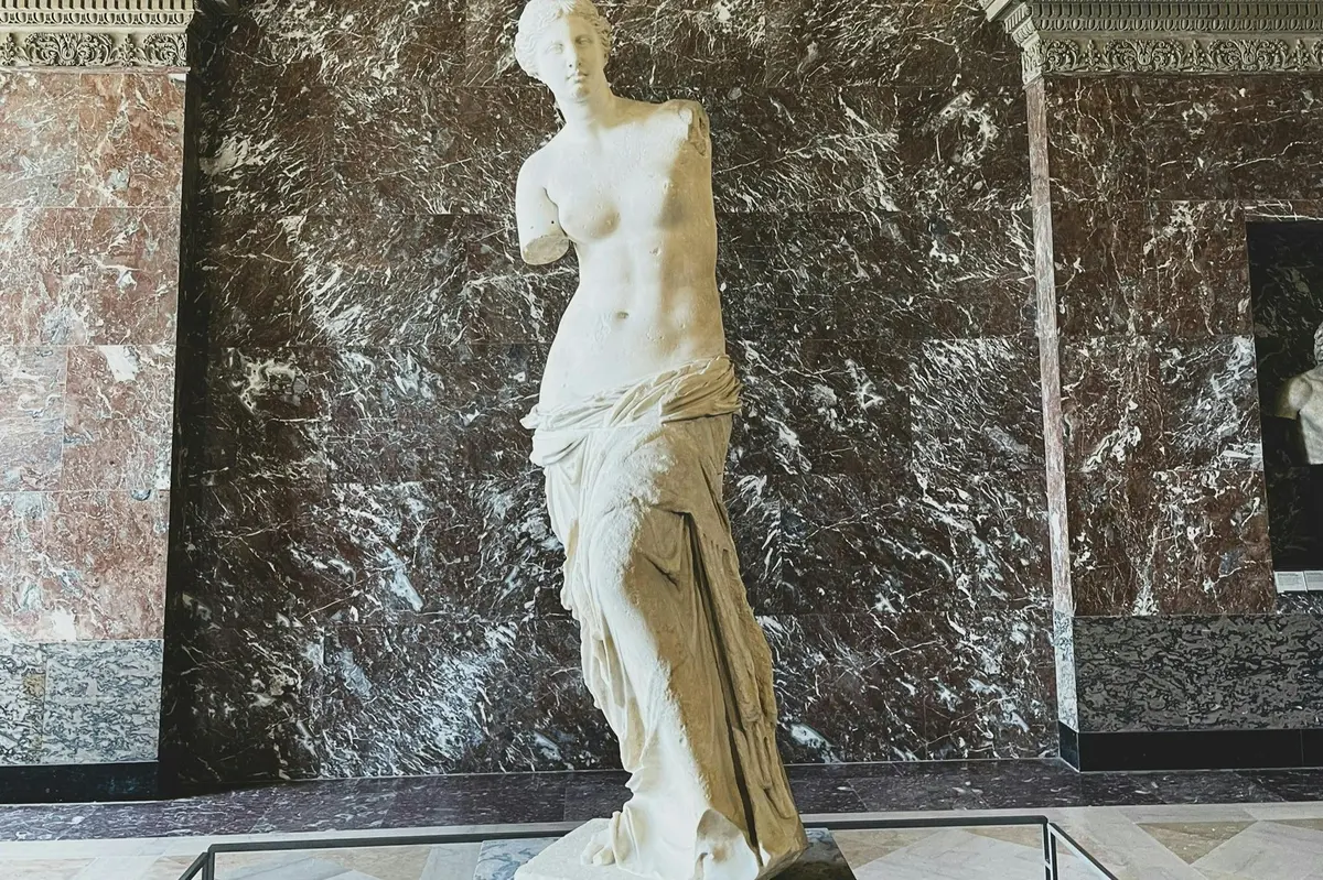 Statue en marbre d'une figure classique sans bras, drapée de tissu, exposée contre un mur de marbre sombre.