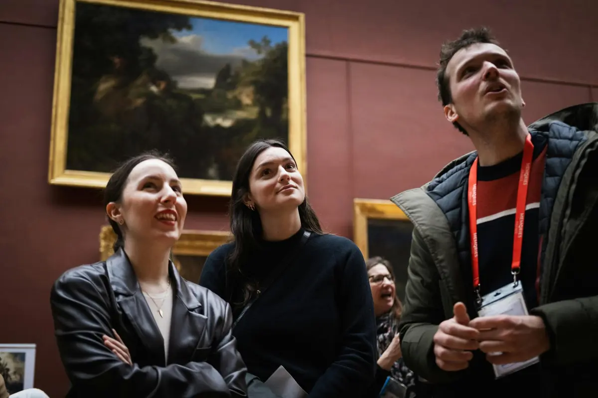 Two women standing in a museum, smiling and looking upward, with framed artwork displayed on the wall behind them.