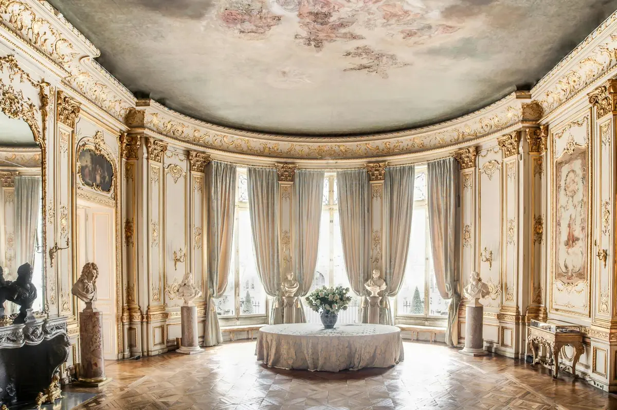 Rotonde élégante aux détails dorés, hautes fenêtres drapées de rideaux, statues, table ronde avec un centre de table floral.