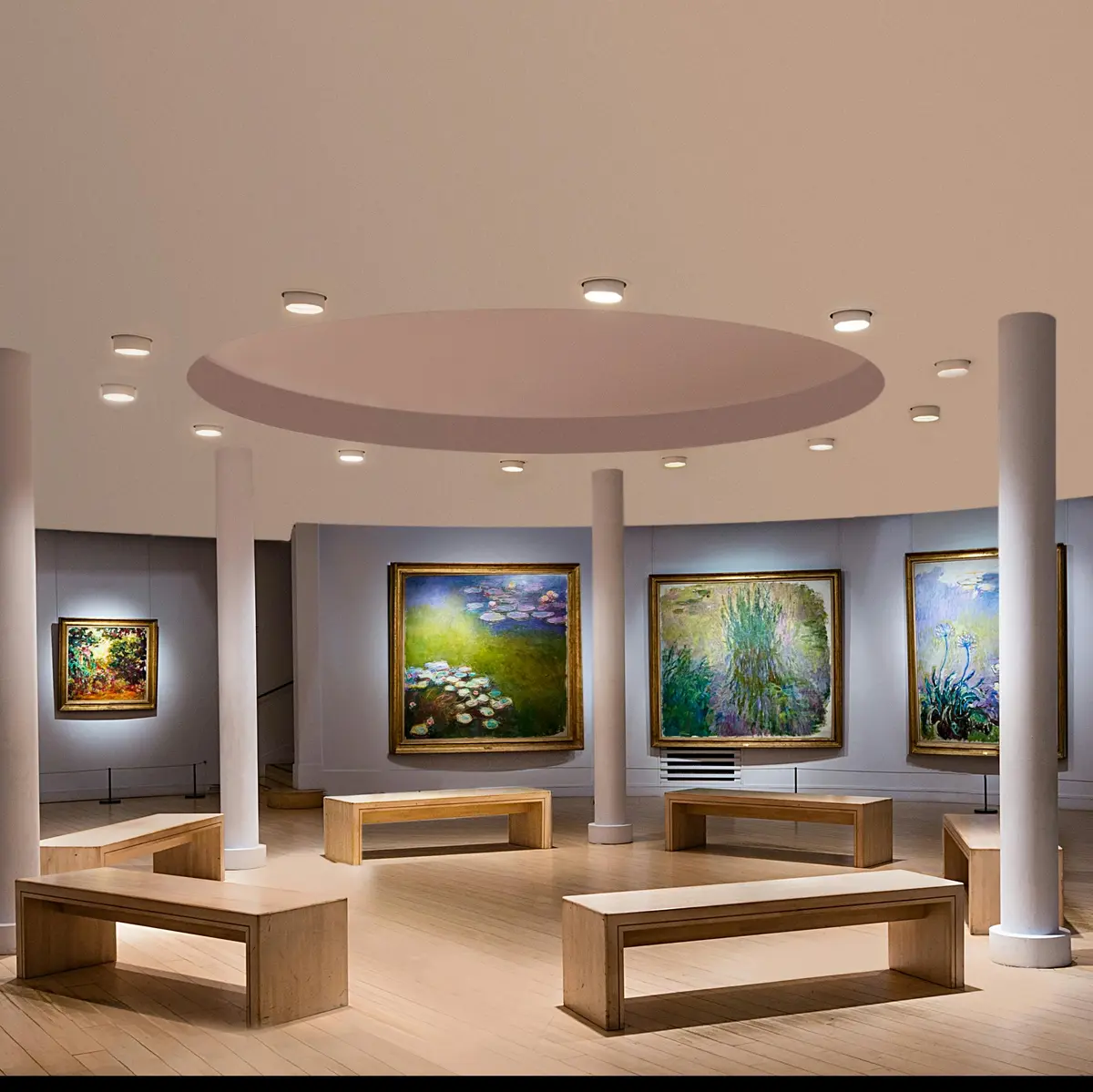 Intérieur d'une galerie d'art avec des bancs en bois et plusieurs grands tableaux encadrés exposés sur des murs gris incurvés sous des plafonniers.