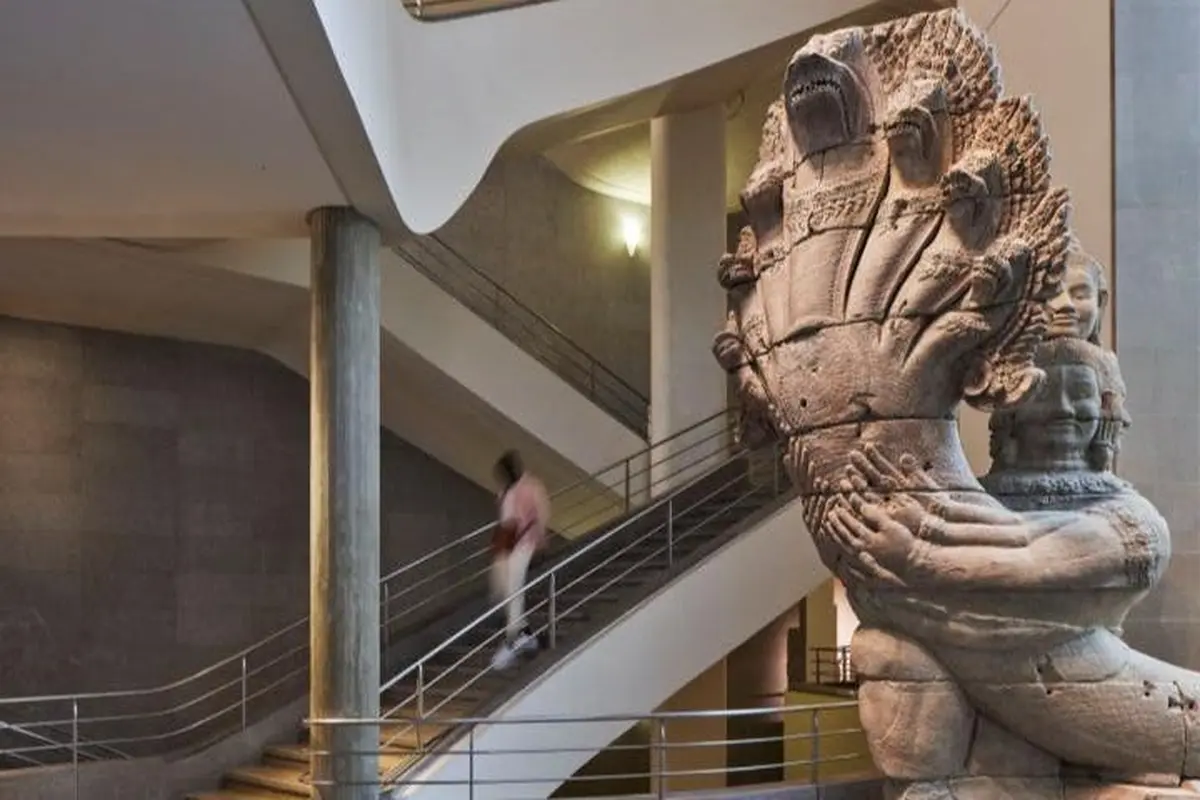 Grande sculpture en pierre représentant un serpent à plusieurs têtes et une figure humaine dans un musée, avec un escalier à l'arrière-plan.