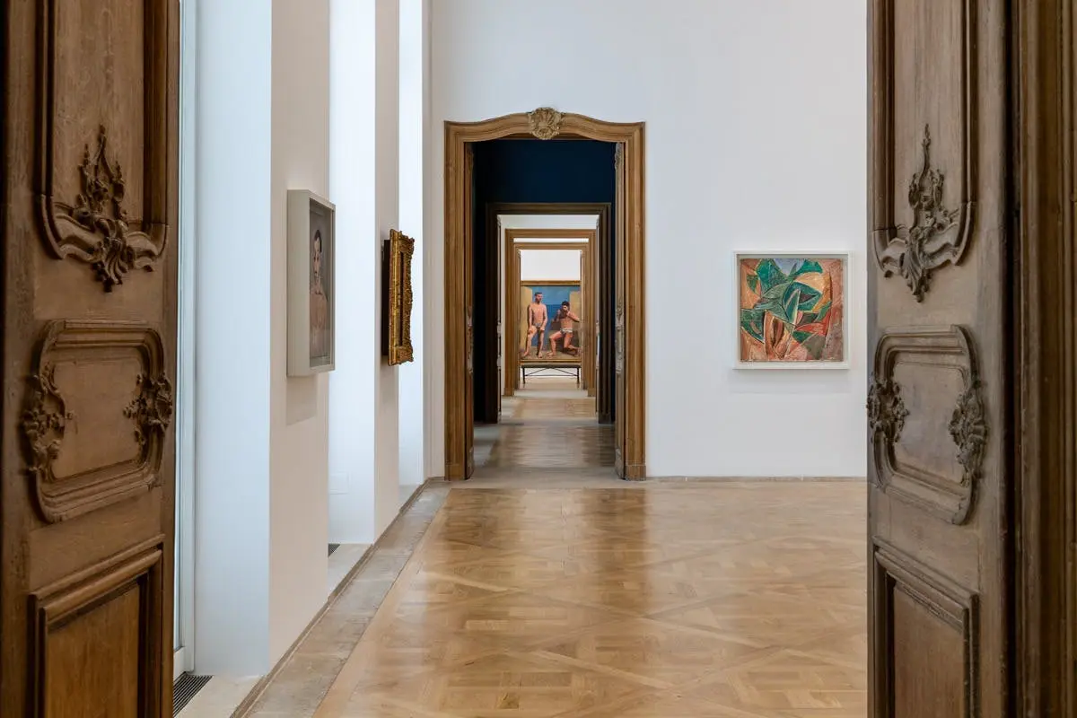 Un couloir de galerie avec des planchers en bois, des murs blancs, des œuvres d'art encadrées et une série de portes ouvertes créant une profondeur visuelle.