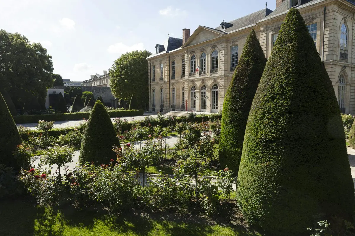 Jardin élégant avec des buissons et des parterres de fleurs soignés devant un grand bâtiment historique aux fenêtres cintrées.