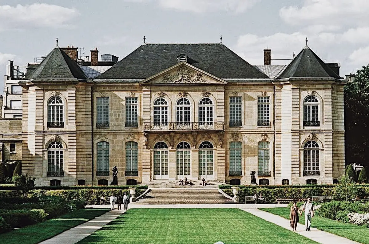 Un grand bâtiment historique avec des fenêtres symétriques et un balcon central, situé derrière une pelouse entretenue où des gens se promènent.