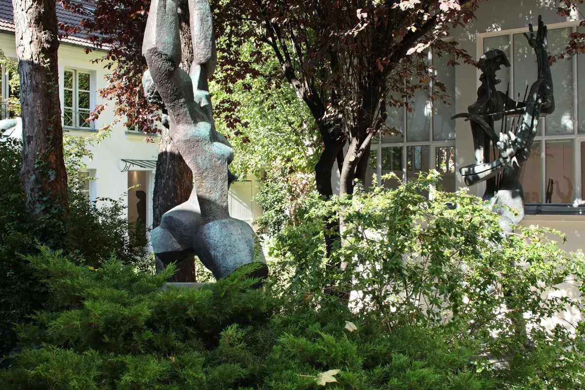 Sculpture en bronze représentant un torse entouré de feuillages et d'arbres verdoyants, avec un bâtiment et une grande fenêtre en arrière-plan.