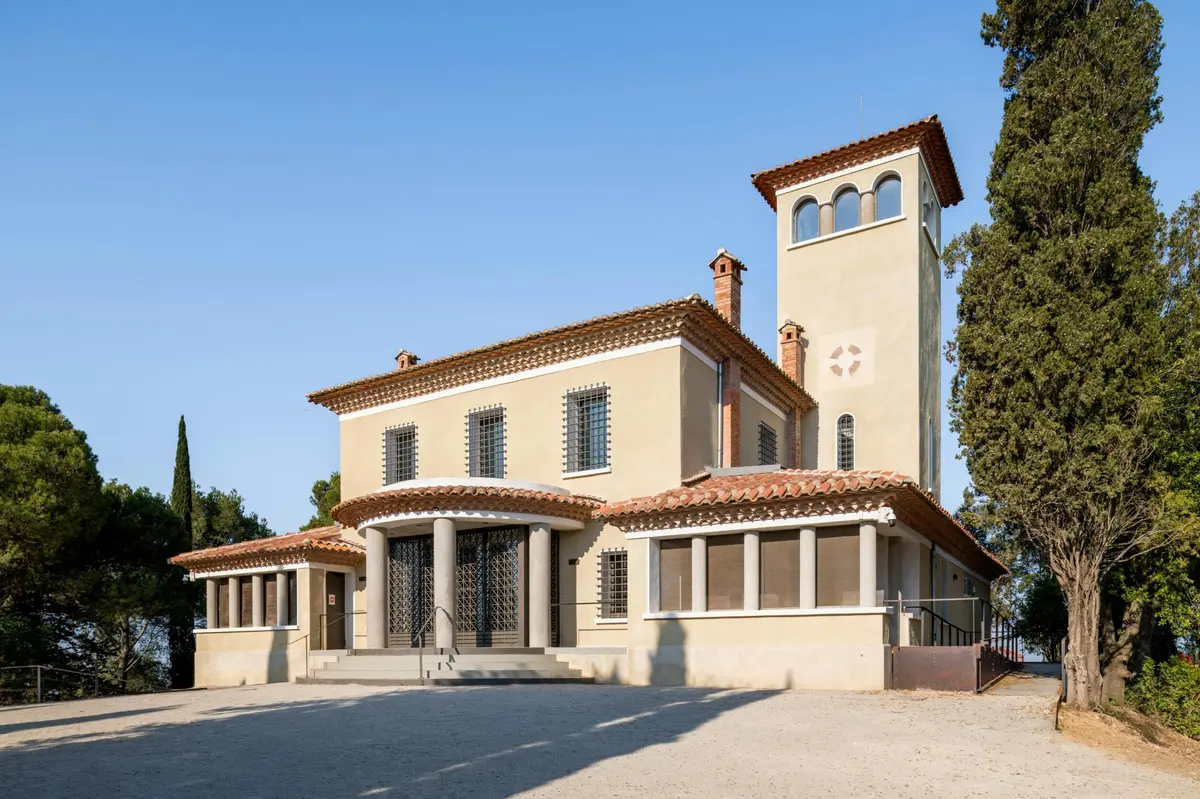 Un bâtiment beige de style méditerranéen avec une tour, un toit de tuiles rouges, des fenêtres cintrées et une entrée à colonnes sous un ciel clair.