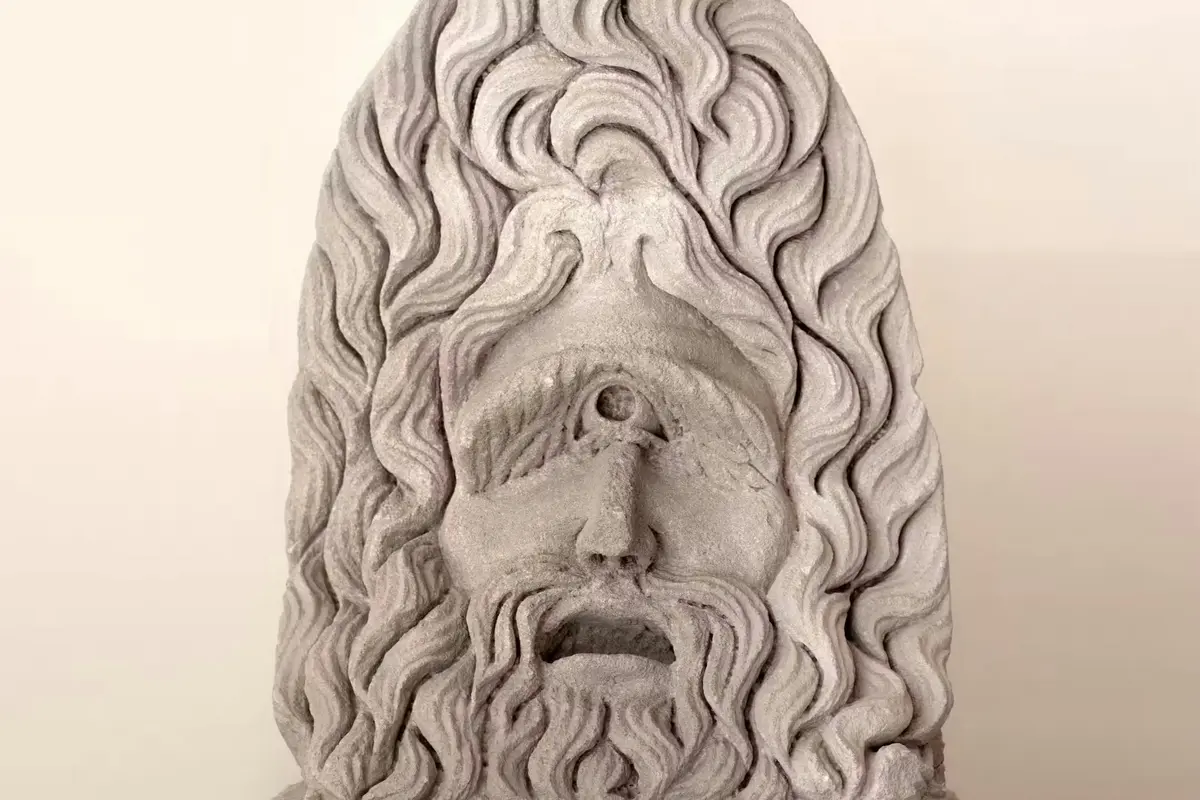 Sculpture en pierre représentant un visage barbu avec un œil unique au centre du front, encadré par des cheveux ondulés.