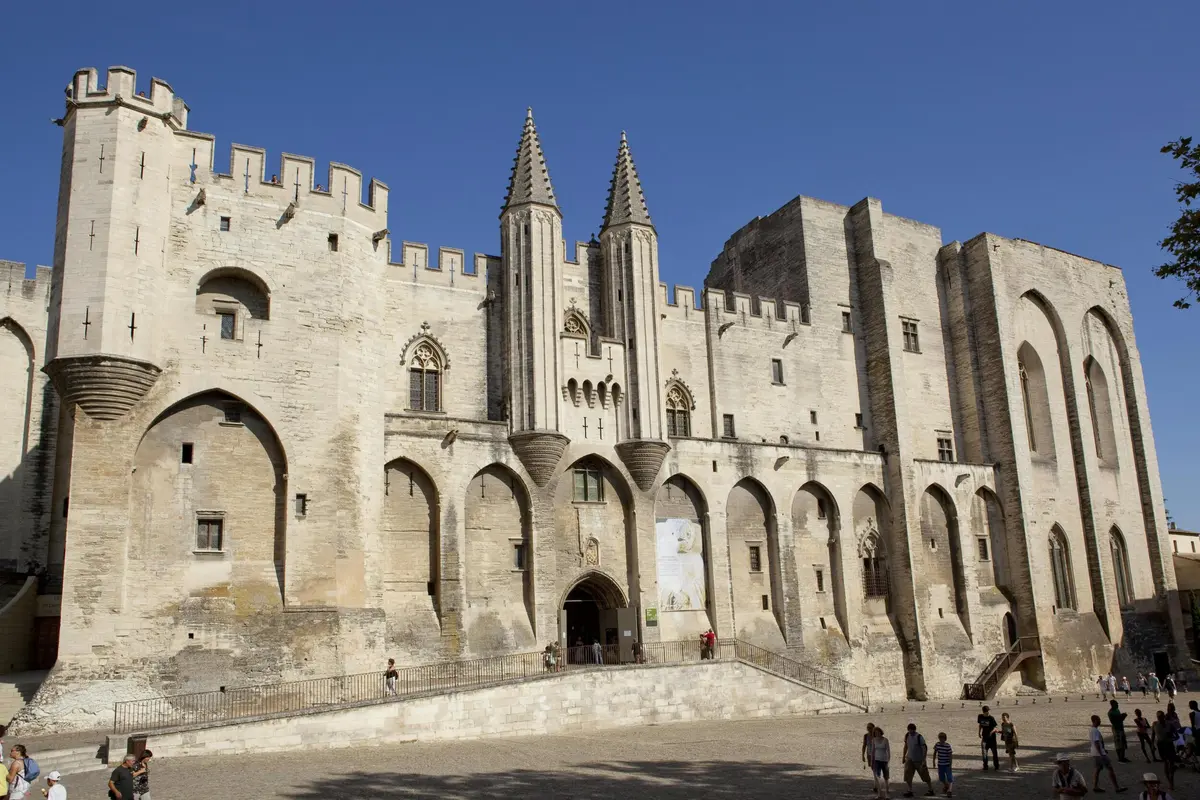 Bâtiment historique en pierre avec de hautes tours, des entrées voûtées et un créneau. Quelques personnes marchent au premier plan. Le ciel est dégagé.