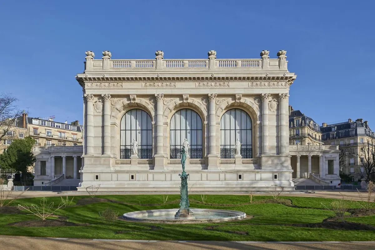 Bâtiment orné de grandes fenêtres cintrées, de statues en façade et d'une fontaine décorative dans un jardin sous un ciel bleu clair.