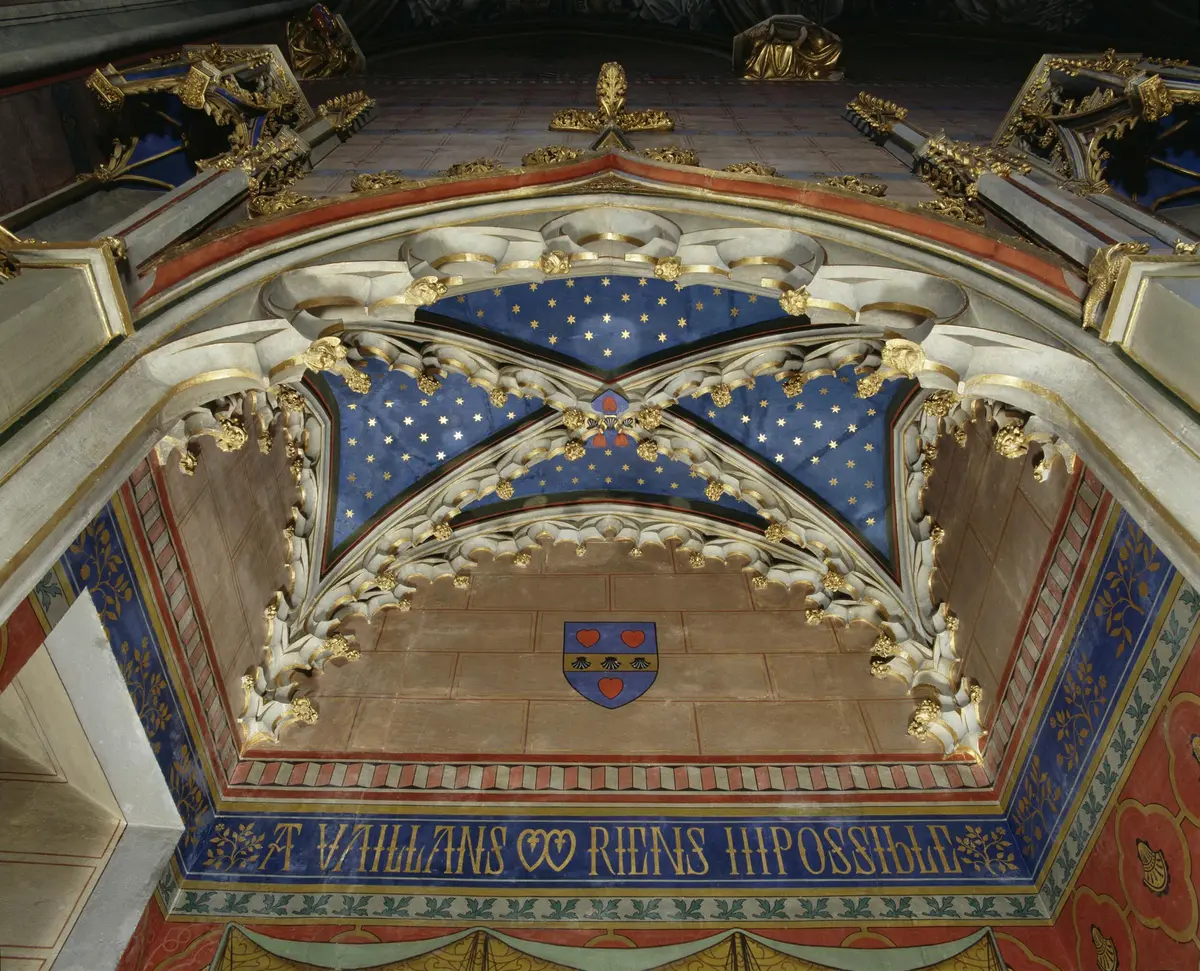 Un plafond gothique orné de touches d'or, un panneau de ciel étoilé bleu, un écusson avec des fleurs de lys et une inscription latine au-dessous.