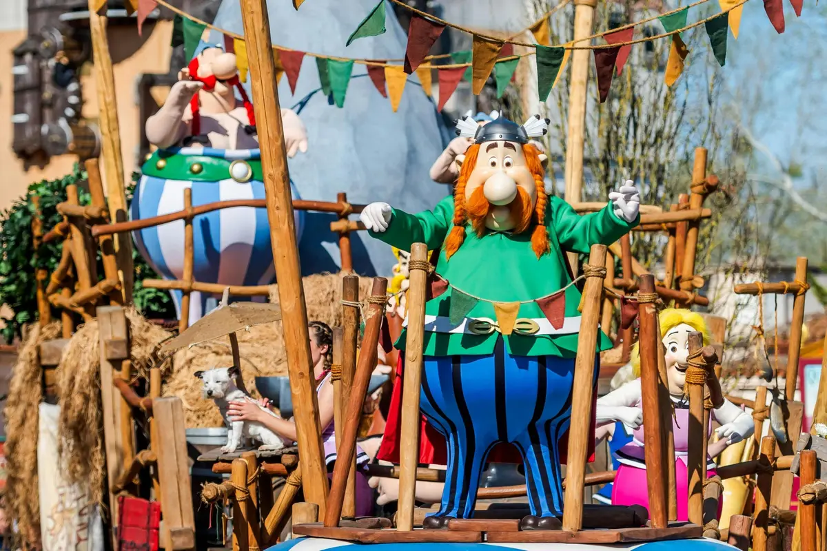 Des personnages de dessins animés hauts en couleur dans un décor festif, avec des guirlandes de drapeaux et des structures en bois, évoquant une scène de défilé.