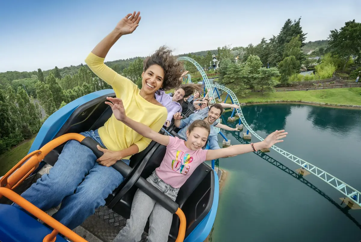 Des personnes profitant d'un tour de montagnes russes, souriant et agitant les bras, avec une vue pittoresque d'arbres et d'eau en arrière-plan.