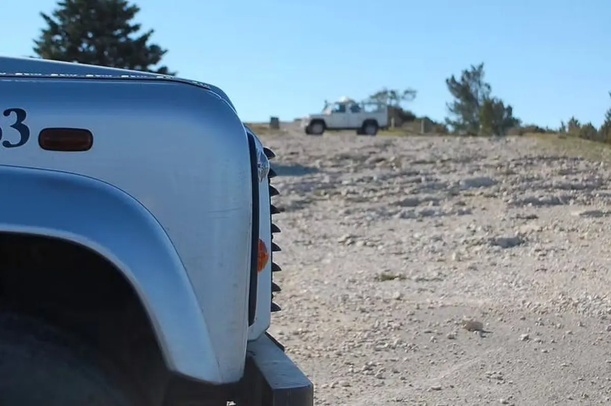 Vue frontale d'un véhicule argenté sur un terrain accidenté et d'un véhicule blanc en arrière-plan, sous un ciel bleu clair.