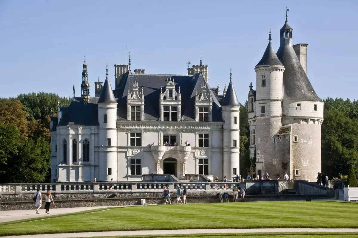 Un grand château de pierre avec de multiples tours et flèches, entouré d'une pelouse bien entretenue. Des personnes marchent près de l'entrée.