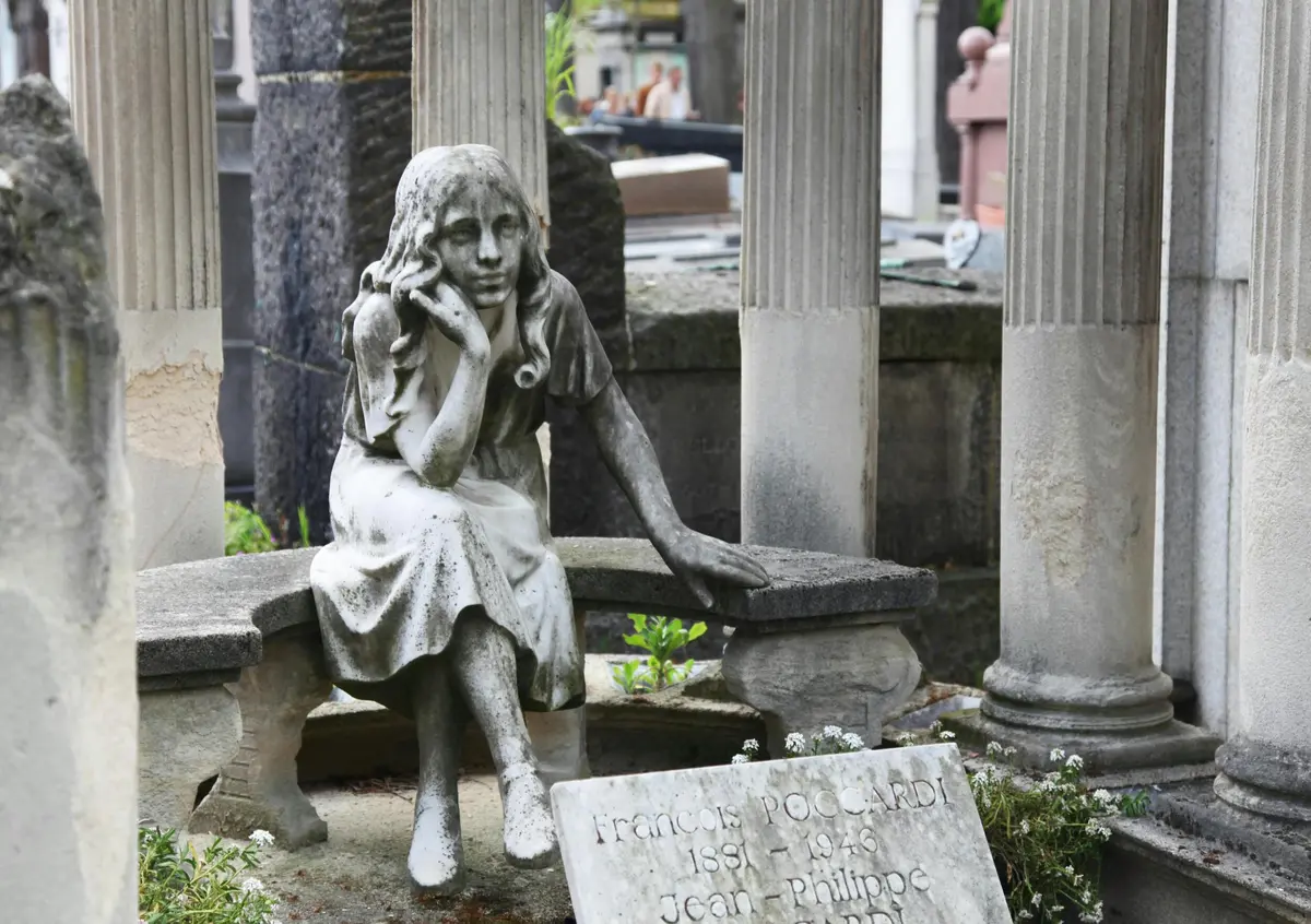 Statue en pierre d'une femme assise, s'appuyant sur une main, entourée de pierres tombales et de colonnes dans un cimetière.