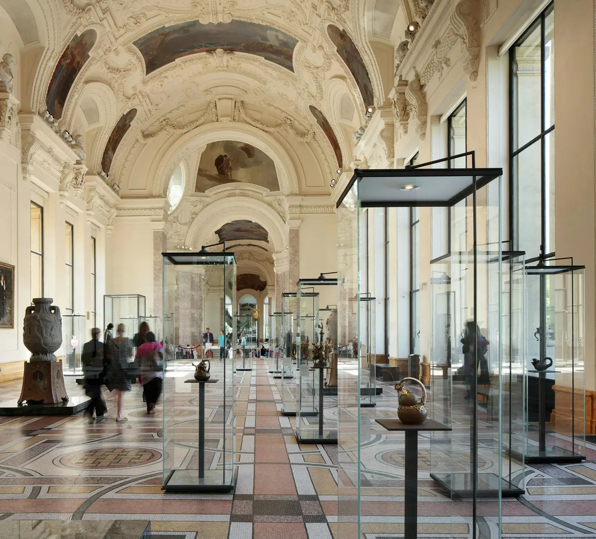 Museum hallway with ornate ceiling and glass display cases containing artifacts. A few visitors walk through the corridor.
