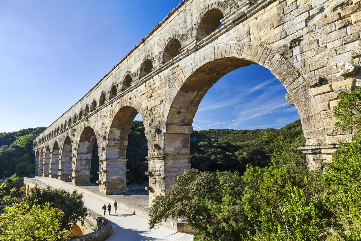 Ancien aqueduc en pierre avec de grandes arches sur fond de ciel bleu et de paysage verdoyant. Trois personnes marchent en contrebas.