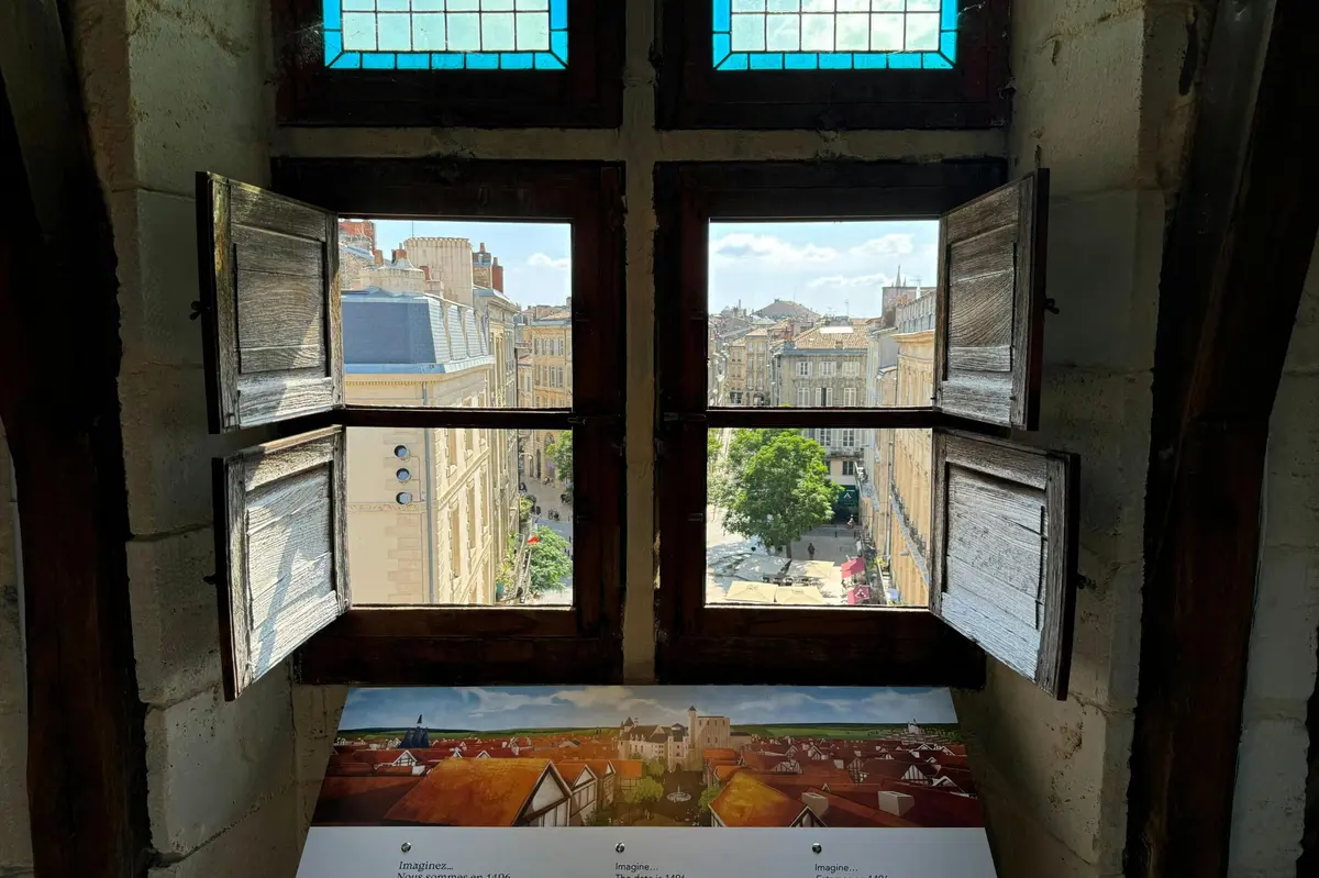 Open wooden shutters reveal a cityscape of historic buildings and streets under a blue sky with scattered clouds.