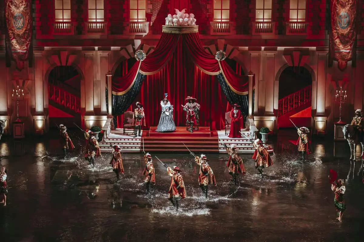 Une représentation théâtrale mettant en scène des comédiens en costumes d'époque, une reine et des gardes sur scène, tandis que d'autres s'éclaboussent d'eau en contrebas.